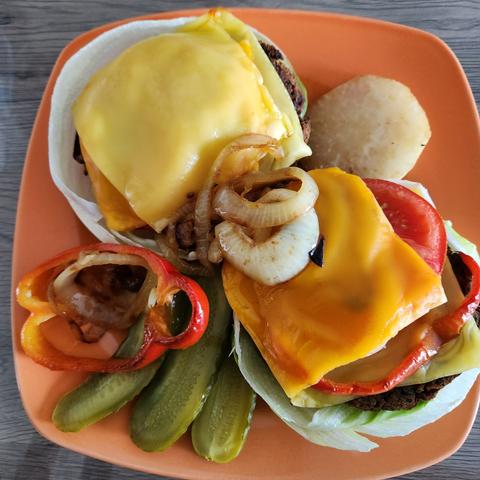 Rote-Bete-Burger auf einen organefarbenen Teller angerichtet. Garnitur Gewürzgurken,  gebratene Zwiebeln und roter, gebratener Paprika. Es sind 2 Burger auf einem Salatblatt gebettet und mit veganen Käse überbacken. Glutenfrei und vegan.