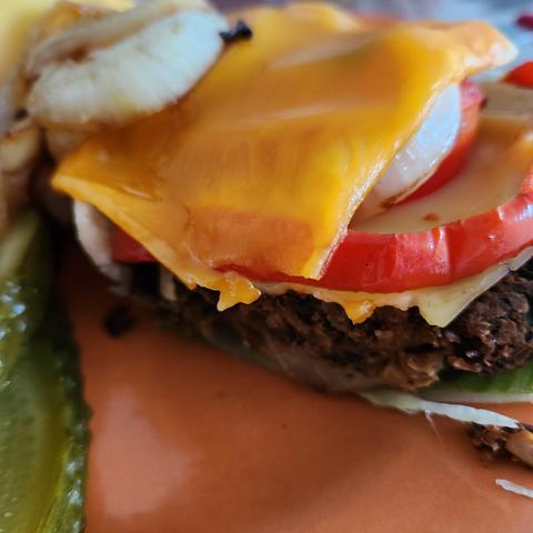 Rote-Bete-Burger auf einen organefarbenen Teller angerichtet. Garnitur Gewürzgurken,  gebratene Zwiebeln und roter, gebratener Paprika. 1 Burger auf einem Salatblatt gebettet, Nahaufnahme, angeschnitten und mit veganen Käse überbacken. Glutenfrei und vegan.