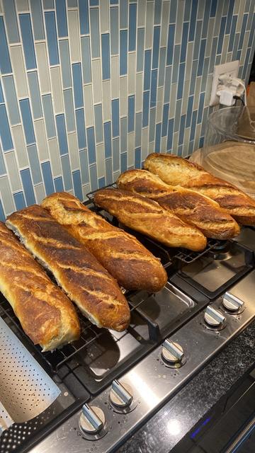 Six golden brown home made baguettes sitting on cooling racks on top of a gas stove.