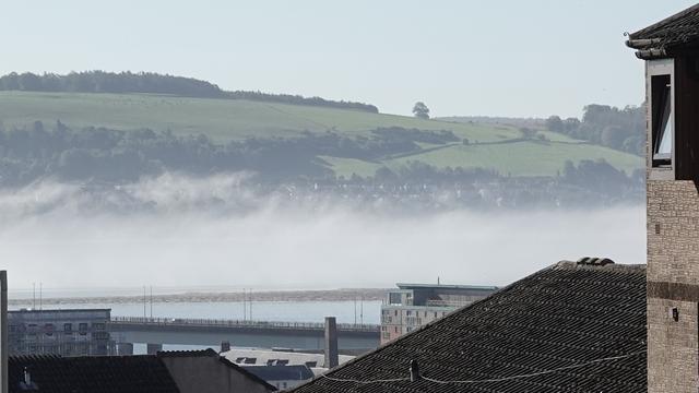 Looking across a haar covered River Tay towards the green hills of Fife. In the foreground are various old Dundee buildings. The Tay Road Bridge is visible, and a sandbank in the river beyond.
