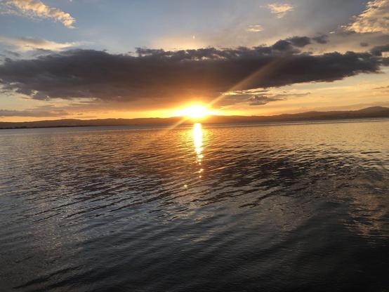 photo: sunset over a lake rimmed with low mountains, a stretch of cloud is above the sun, framing it against the mountain horizon.