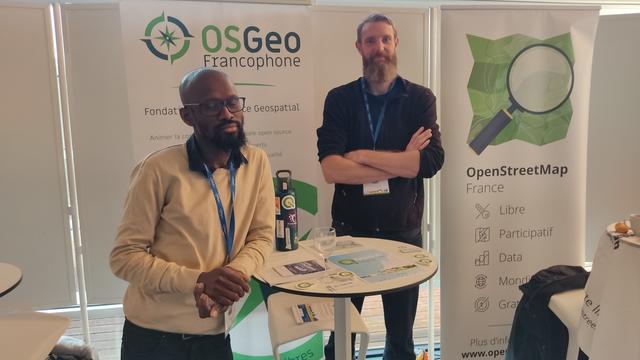 deux hommes barbus se trouve derrière une table de type mange-debout avec en arrière plan les kakémonos de l'association osgeo-fr et de l'association OSM France