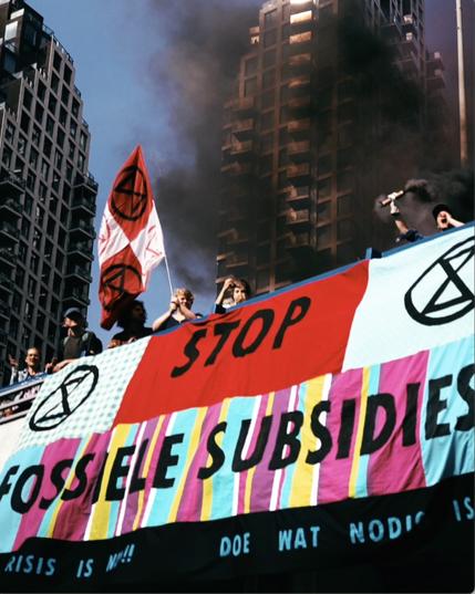 Beeld van een aantal rebellen met rookpotten, XR vlaggen en een groot spandoek met daarop XR logo’s en de tekst ‘stop fossiele subsidies’