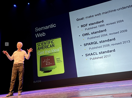 Ian Horrocks standing on the big stage at his Semantics 2024 keynote. In the background a slide is displayed with the cover of the famous Semantic Web Article from Scientific American (back from 2001) and a few dates for the SemWeb standards such as RDF, OWL, SPARQL, and SHACL.