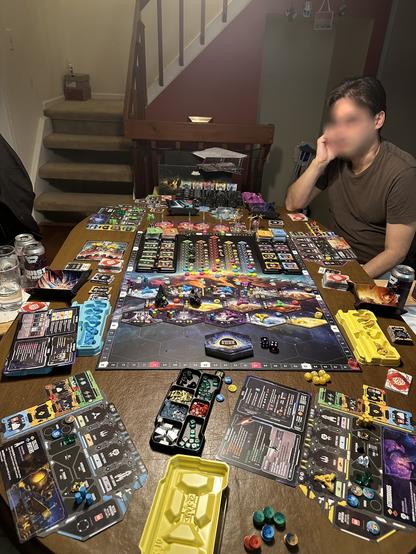 A complex space themed board game is arranged on a table. It takes up all of the surface space. On the board you can see planet tiles, plastic ship miniatures, larger detailed raider minis, large development minis, and an array of cards and fancy resource tokens for ice, credits, titanium, nanocarbon, and energy. While there are four placemats set up, only one player is visible in the photo and his face is blurred for privacy.