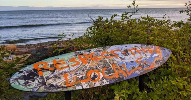 A painted old surfboard, made into a mounted sign beside the beach & in front of the Pacific Ocean. It says, in bright orange paint:
RESPECT THE LOCALS