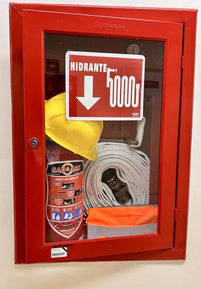 A red fire safety cabinet containing a fire extinguisher, fire hose, a yellow hard hat, and an orange safety garment. A sign labeled "HIDRANTE" with an arrow pointing down is visible on the front.