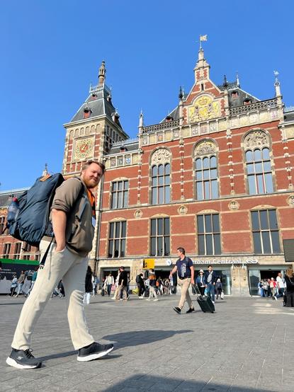 Markus (@markus@skvip.lol) står framføre Amsterdam Centraal-stasjon. Han har på seg ein stor blå sekk. Han har ljosebrune buksar, ein brun genser, og sorte sko med kvite sålar.
Det er blå himmel.

Amsterdam Centraal er eit stort gamaldags bygg. Raud stein. Brune og kvite aksentar.

Det er mange folk mellom Markus og stasjonen.