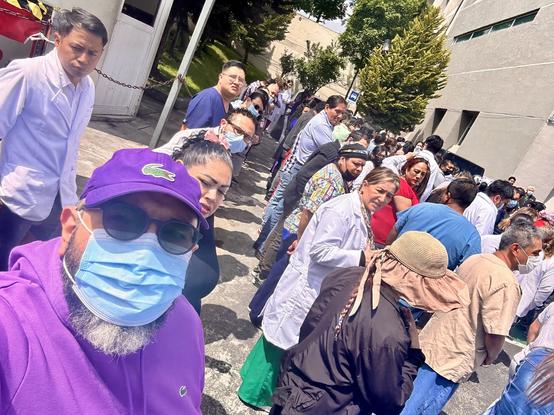 A large group of people is lined up outside a building, many wearing masks. The scene captures a diverse crowd, including individuals in medical attire and casual clothing, waiting for simulated earthquake on a sunny day.