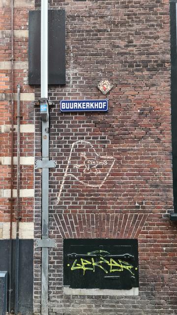 A simple 2d cream white chalk rendition of a flag with a skull and bones on a grimey red brick wall next to a square drainpipe and below a blue with with letters streetname sign reading buurkerkhof (Neighbor cemetary).