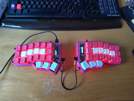 My finished Corne keyboard on my desk. It is a 42 key split keyboard with a purple case. The keyscaps are pink except the white home row and the light blue thumb keys. All keys got a red lighting and under the keyboard I'd a red underflow visible. Each side of the keyboard has a little display. One shows the Corner logo, the other some layer data.
In the background you can see part of my old Cherry keyboard with customised keycsps