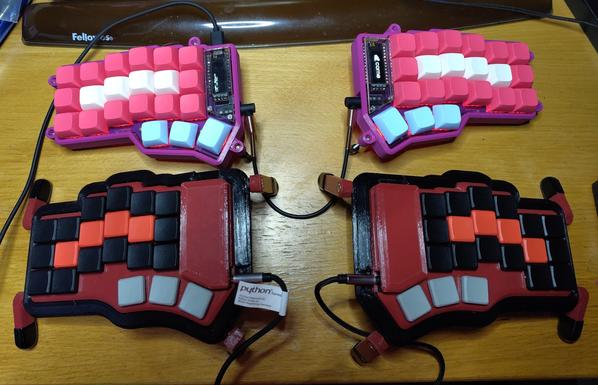 On top is the Corner keyboard mentioned in the other image. In the lower part you can see my Cantor keyboard. The key layout is similar but slightly more stretched. The keycaps are mainly black with a red home row and grey thumb keys. It's got neither lighting nor displays. The case is black and red.