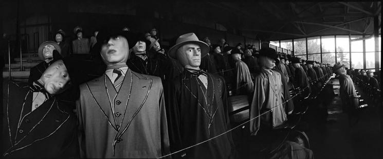 Black and white Widelux panoramic 35mm photo by Jeff Bridges. The wide photo shows a closeup of several rows of crude dummies — mostly men — dressed in 1930’s style suits and hats. They are posed standing in rows of seats in what looks like a covered race track stand. Likely for a simulated crowd scene of a movie.