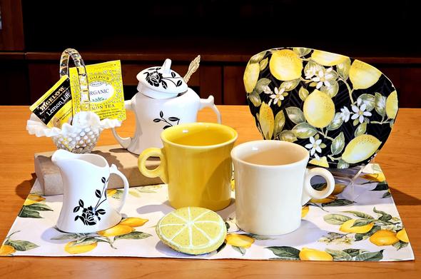 A tea tableau in black and yellow with lemon-themed cozy, placemat, and a rock painted to look like a lemon slice. The mugs are Fiestaware in two shades of yellow, and there is a white cream & sugar set with black scrollwork. The teas are Bigelow Lemon Lift and St Dalfour Lemon, two packets in a white hobnail milk glass basket.