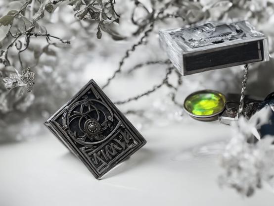 Close-up of a tiny, ornate silver book-shaped locket, with intricate details and a green gem, surrounded by other delicate accessories, against a wintry, monochromatic background. Macro photography style.