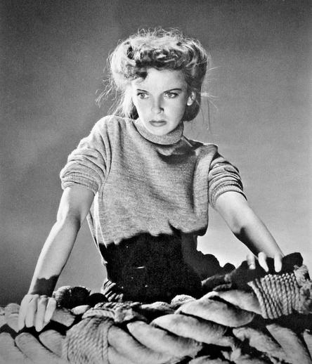 Studio photo of Lupino in simple sweater with sleeves pushed up, leaning on pile of very large rope.