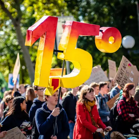 Foto eines Klima-Streiks. Eine Person hält 1.5° (3D Modell) nach oben. Die Zahl steht für das internationale Abkommen von Paris, die mensch-gemachte Erwärmung unseres Planeten auf diese durchschnittliche Erwärmung zu begrenzen. Die Zahlen in Gelb, die nach Rot wechseln als Symbol für die Dringlichkeit der Aufgabe.