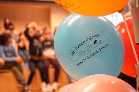Das Bild zeigt Luftballons mit dem Logo der Daten-Füchse. Im Hintergrund sitzen Kinder (das Bild ist dort unscharf).