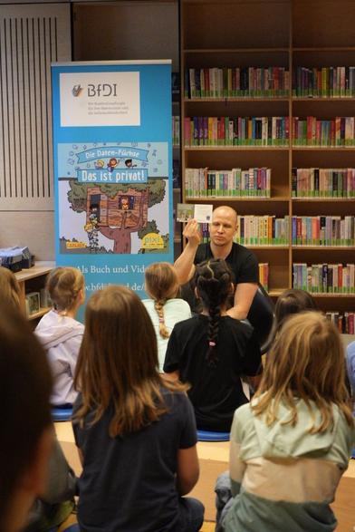 Das Bild zeigt einen BfDI-Mitarbeiter, der Kindern aus einem Pixi-Buch vorliest.