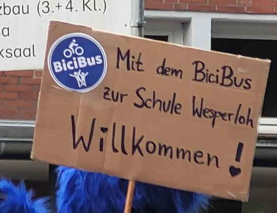 Ein braunes Pappschild. Darauf ein blauer, runder Aufkleber mit einem weißen Fahrradpiktogramm, einem kinderpiktogramm und dem Wort bicibus. Daneben schwarzer text: " mit dem bicibus zur Schule Wesperloh. Willkommen!"
Das Schild wird von einem blauen, flauschigen Monster gehalten.