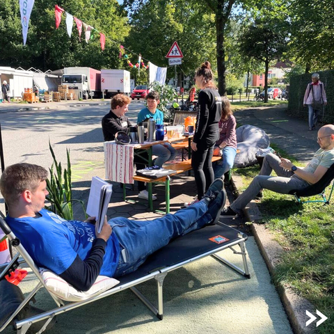 Man sieht einen besetzten Parkplatz, auf dem Menschen auf Liegestühlen entspannen oder an einem Tisch Kaffee trinken.