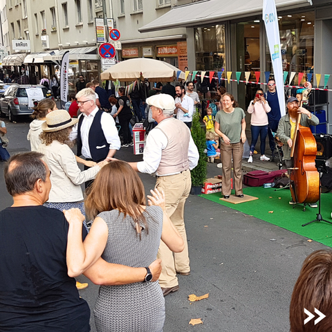 Man sieht eine bunt geschmückte Straße auf der Menschen tanzen. Wo normalerweise ein Parkplatz wäre, spielt eine Liveband Musik.