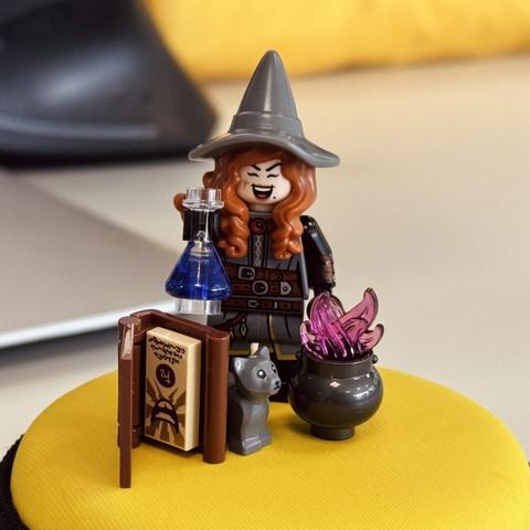 Minifigura de Lego de Tasha, de Dungeons And Dragons. Está sobre un estuche amarillo y redondo de auriculares. Sale ella con su sombrero, su melena castaña, la pata de pollo en la mejilla y el lunar. En la mano tiene un frasco con una poción y a los pies está el Demonomicón (libro), un gato y, claro, el caldero, del que salen como unas llamas moradas.