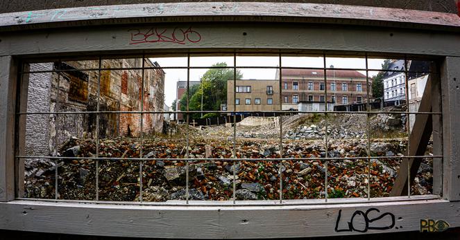 A small-paned window without glass, divided by rebar, fills the entire color photograph. Behind the window opens up a demolition site with lots of old red bricks. In the background, on the other side of the city quarter, a number of business premises on the other side of the next city street.
🇸🇯Et smårutet vindu, delt opp med armeringsjern, uten glass fyller hele fargefotografiet. Bak vinduet åpner det seg en rivningstomt med mye gammel teglstein. I bakgrunnen, på andre siden av bykvartalet en rekke forretningsgårder på andre siden av neste bygate.