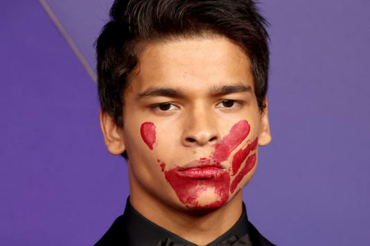 An image of a young Native American man, with short black hair, and dark eyes, with a red hand print on his face. He has a very serious expression on his face, and is wearing a black suit, dress shirt, and tie.