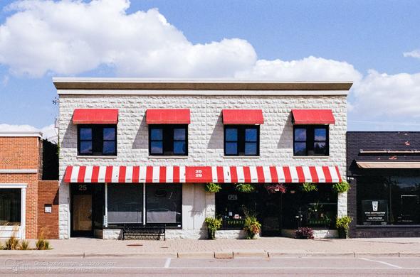 A photograph of a white painted, two-story block building, in the summer sun beneath a partly-cloudy sky. It has red awnings over four windows on the second story, and a red-and-white-striped awning along the shops on the bottom floor. It is flanked by a red brick neighboring building on the left, and a black-painted brick building on the right.