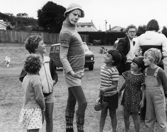 The image portrays an outdoor scene at Ben Burn Park, Karori. A person dressed as a clown or mime is standing in the center of the frame and appears to be entertaining several children who are gathered around him/her. The group consists of six young kids with various expressions ranging from laughter to curiosity.

In the background, there's another adult male facing away from the camera, seemingly engaged in conversation with someone off-frame. Onlookers can also see multiple vintage vehicles parked on a grassy field and some trees that provide shade over parts of the scene. The image is presented in black-and-white, which gives it an old-fashioned or historical feel.

Additional information about this photo indicates that it captures a moment from Ben Burn Park during a free outdoor concert organized by R&B band Rough Justice at Campbell Street, Karori. This event was sponsored by Radio Windy, one of the first private radio stations operating in Wellington. The photographer is credited to A... https://images.loener.nl/AnsWestra/full/6690/6690f4813c8dcd5ca3a70537.jpg
