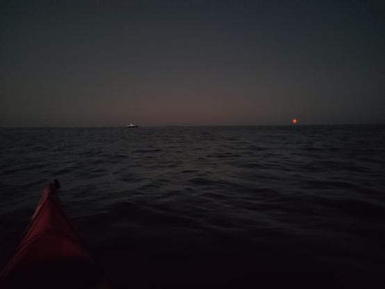 A red moon rising out of a dark sea. The point of a red kayak is in the foreground. A white boat approaches the moon in the background