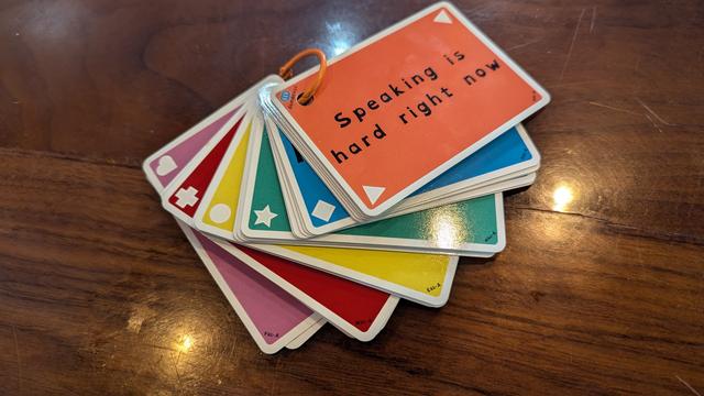 A ring-bound series of colorful cards is fanned out on a dark wood countertop. The top card is orange and reads "Speaking is hard right now." It has white triangles in two opposite corners. Visible beneath are cards in blue, green, yellow, red, and pink; with diamond, star, circle, plus, and heart symbols respectively.
