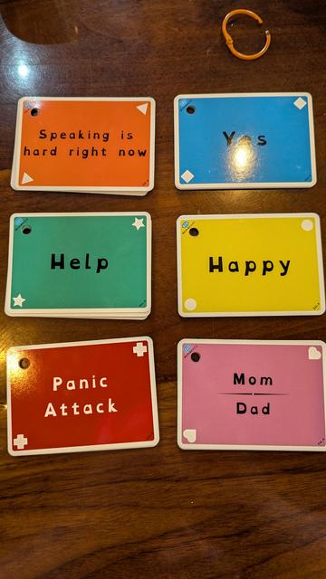 Six small piles of colorful cards are arranged in a dark wood countertop. The top cards are the same from each section of the set as in the previous picture. Starting at the upper left: an orange card with triangles in its opposing corners reads "Speaking is hard right now." A blue card with diamonds in its corners reads "Yes" (though a glare obscures the 'e' somewhat). A green card bearing stars reads "Help." A yellow card bearing Circles reads "Happy." A red card bearing plus signs and white text reads "Panic Attack." Finally, a pink card bearing hearts reads "Mom / Dad."
