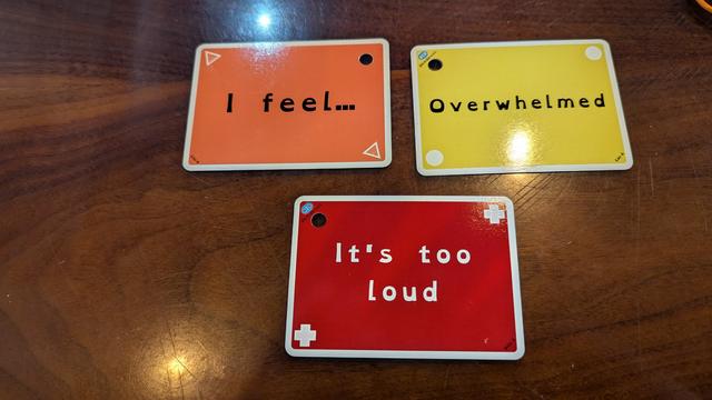 Three cards from among the set are arranged on a dark wood countertop: An orange card bearing triangles flipped to its backside, a yellow card bearing circles, and a red card bearing plus signs form the sequence "I feel overwhelmed. It's too loud."