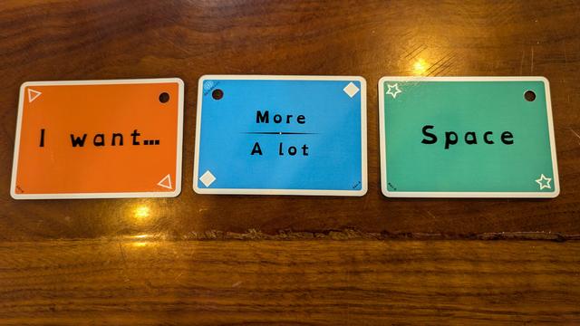Three cards from among the set are arranged on a dark wood countertop: An orange card bearing triangles flipped to its backside, a blue card bearing diamonds, and a green card bearing stars flipped to its backside form the sequence "I want (more / a lot) space."