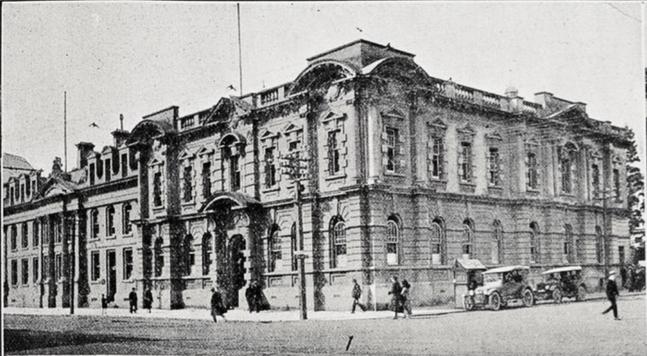 Black-and-white photo: [Detail from] Some of Wellington’s public buildings and colleges. 1922. Description: Detail from the photo montage, showing the Magistrate’s Court on a street corner, with two cars (possibly taxis) parked on the kerb and pedestrians walking on the road and footpaths (BrE pavements, AmE sidewalks). The court building is two storeys and clad in stone. Citation: Supplement to the Auckland Weekly News, 21 September 1922, p. 39. Auckland Libraries Heritage Collections AWNS-19220921-39-01. https://kura.aucklandlibraries.govt.nz/digital/collection/photos/id/238986