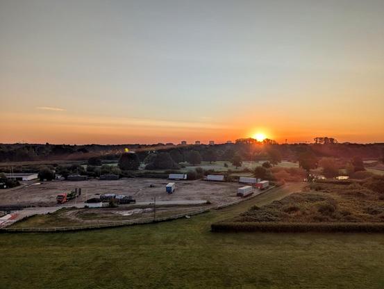 Ein Sonnenaufgang über Hamburg. im Vordergrund ist ein großer, grüner Teil der Trabrennbahn. Mittig im Bild ist ein Teil einer Baustelle mit Fahrzeugen zu sehen. Dahinter ist etwas Wald und leicht nach rechts versetzt scheint die Sonne orange rot durch Bäume hindurch. Der Himmel ist rot gefärbt.