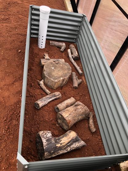 Various sized pieces of wood and a white PVC pipe rest on red soil inside a corrugated garden bed not yet topped off with soil and compost.