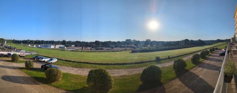 Ein Panorama der Rennbahn hinter dem Hotel. Links am Bildrand sieht man die Ausläufer eines Flohmarktes. Viel grüne Fläche in der Mitte. Bäume im Vordergrund unterbrechen die Wiese. Rechts mittig blendet die Sonne am blauen Himmel.