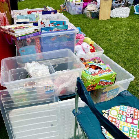 A variety of clear plastic storage boxes filled with toys, books, and other items are arranged on a grassy area. A folding chair with a colorful patterned cushion is visible in the foreground, and additional boxes and items are present in the background.