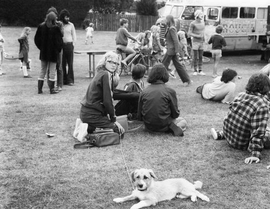 Black and white photograph of people gathered in a park for an outdoor concert by the band Rough Justice. Individuals are seated on grass, benches, or standing around tables with drinks and refreshments laid out before them. A dog is lying peacefully close to one person's feet as they sit cross-legged on the ground. In the background stands another bus labeled "Rough," likely a touring vehicle for the band members.