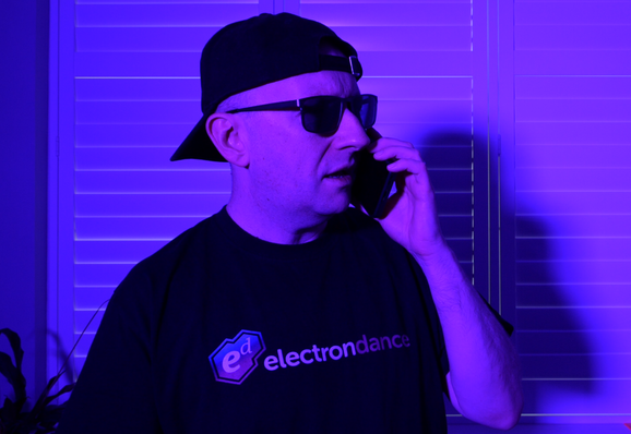 Joel Goodwin aka Electron Dance talking on a phone, backward facing cap, bathed in purple light.
