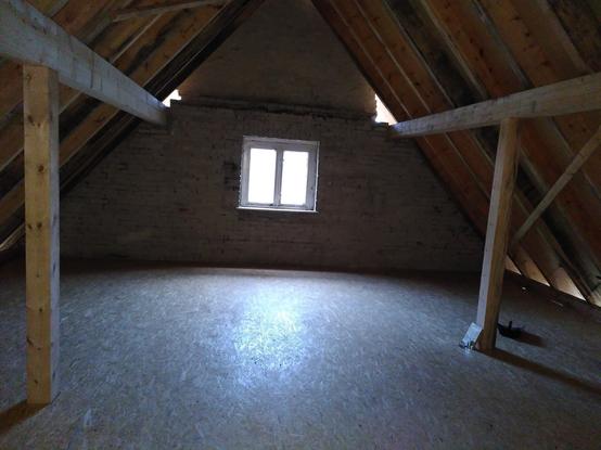 Foto aus dem Inneren eines neu errichteten Dachstuhls aus naturbelassenem Fichtenholz, teils hell, teils vergraut. Links und rechts stehen Holzpfosten, auf denen die Pfetten und Sparren des Dachstuhls liegen, der Boden ist mit OSB-Platten ausgelegt. Durch ein Fenster in der Giebelwand im Zentrum des Bildes fällt Licht in den Raum.