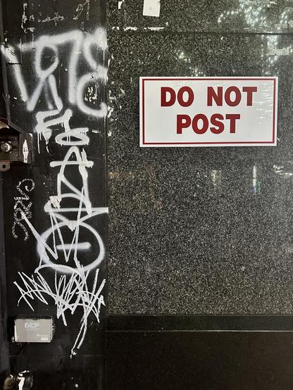 a wall with a bunch of graffiti tags in white, including ANSO in vertical letters with a halo over the A. the right panel is shiny gray granite with a white sign saying DO NOT POST in red letters.