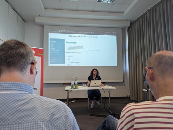 Viviana hält einen Vortrag zum Thema CSS. Sie sitzt vor einer Leinwand an einem weißen Tisch mit ihrem Laptop. Alle warten gespannt.