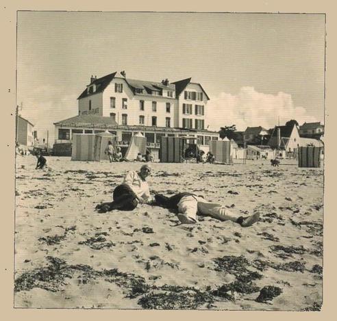 Jacques Tati sits on a beach in Saint-Marc-sur-Mer (he lies down at the same time). He looks at the camera. Another crew member (probably) is resting besides him. We see the hotel from the film in the background. B/w photo.
