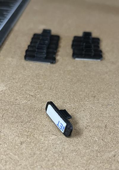 A usb cable clip printed in black filament with 11 more clips behind in two columns.
The clip is a rectangular shape with rounded ends, turned upside down revealing a small rectangle of white foam adhesive.