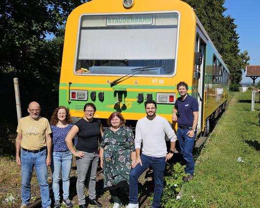 Eine Gruppe Menschen steht vor einem gelben Zug mit grünen Streifen. Oben auf der Abfahrtstafel steht "Staudenbahn". Der Himmel ist blau, man sieht viele Pflanzen um die Bahnlinie
 Die Menschen vor dem Zug sind, von links nach rechts: Reiner Erben, Umweltreferent von Augsburg; ich; Stephanie Schuhknecht, Landtagsabgeordnete für Augsburg; Christine Kamm, Serdar Akin und Deniz Anan, allesamt Stadträte von Augsburg.