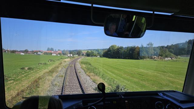 Blick aus dem Fenster beim Schaffner: Schienen in einer grünen Landschaft mit kurz gemähtem Gras, am Horizont einige Häuser. Man sieht keine Stromleitung, daher fährt der Zug derzeit mit Diesel.